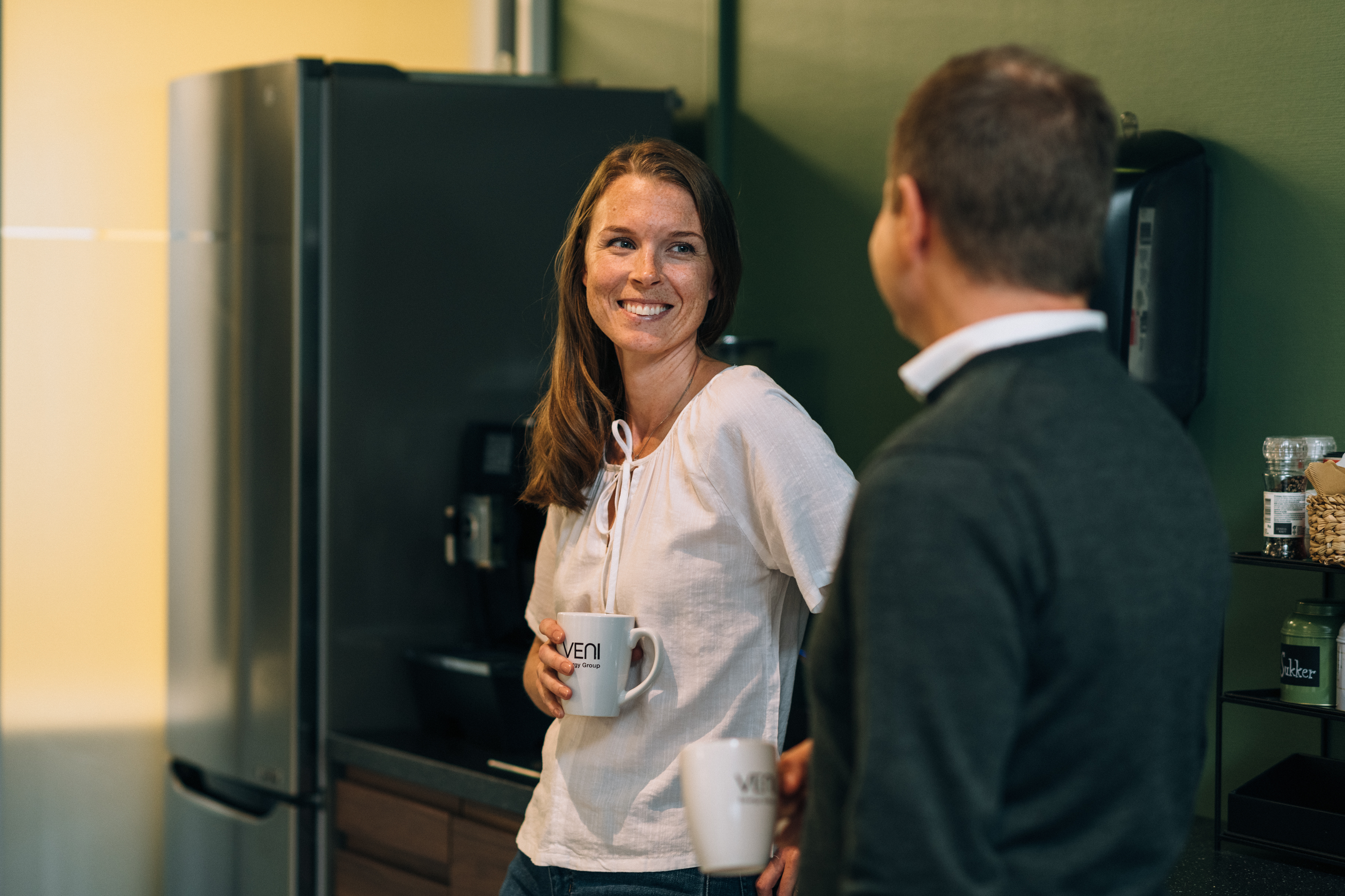 Ansatte har en samtale på kjøkkenet, en kvinne står mot kamera med kaffekopp i hånden, en mann står med ryggen til