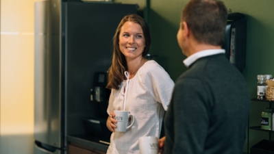 Employees having a conversation in the kitchen, a woman is facing the camera with a coffee cup in her hand, a man is standing with his back to the camera