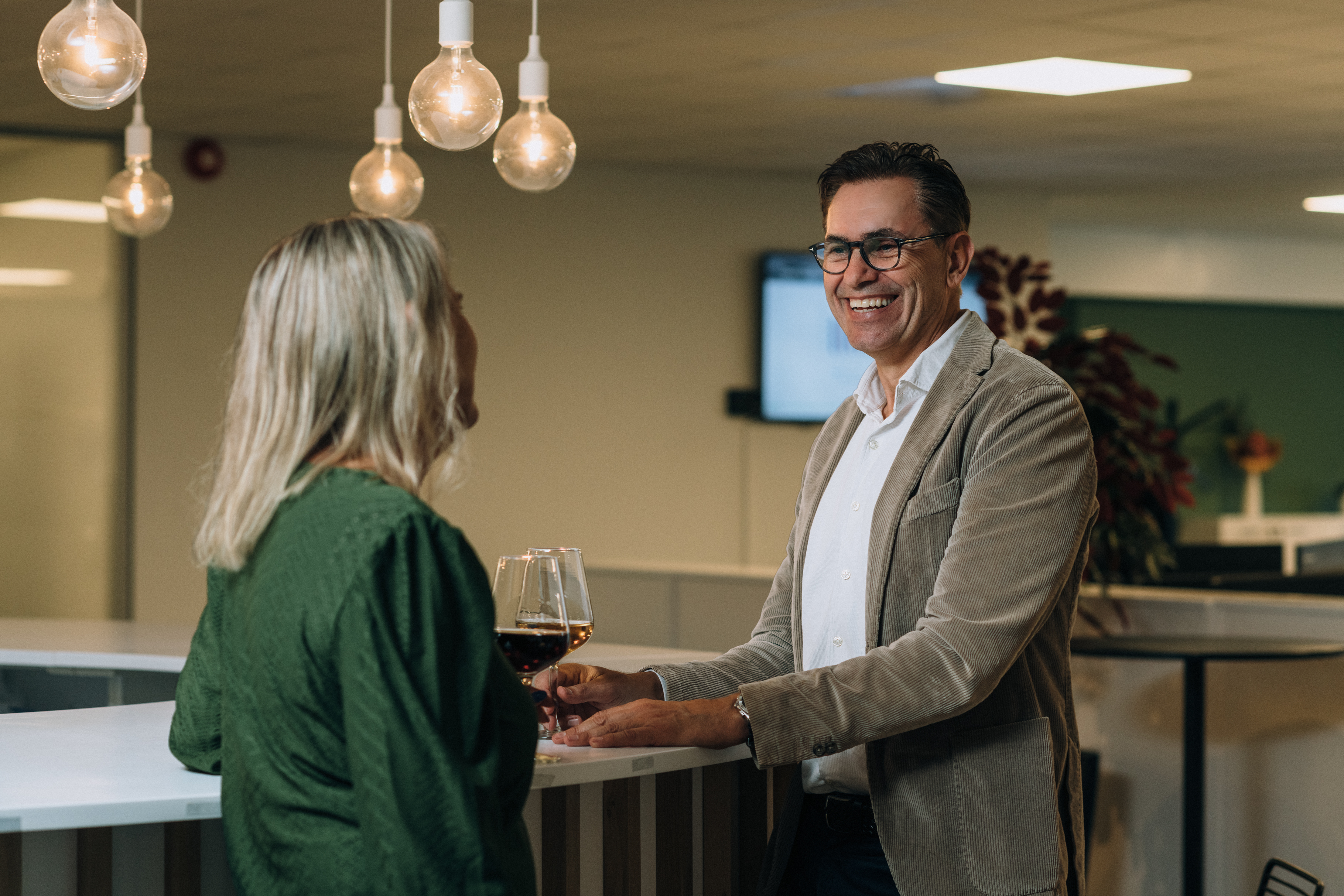 En kvinnelig ansatt sett fra baksiden snakker med ansatt Trond Lutdal i en bar med hengende lyspærer over hodet